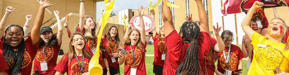 photo of about a dozen 六合图库 students wearing matching marron and gold shirts cheer with their arms raised. With streamers and flags flying, the students exude high energy on a grassy lawn with yellow brick buildings in the background.