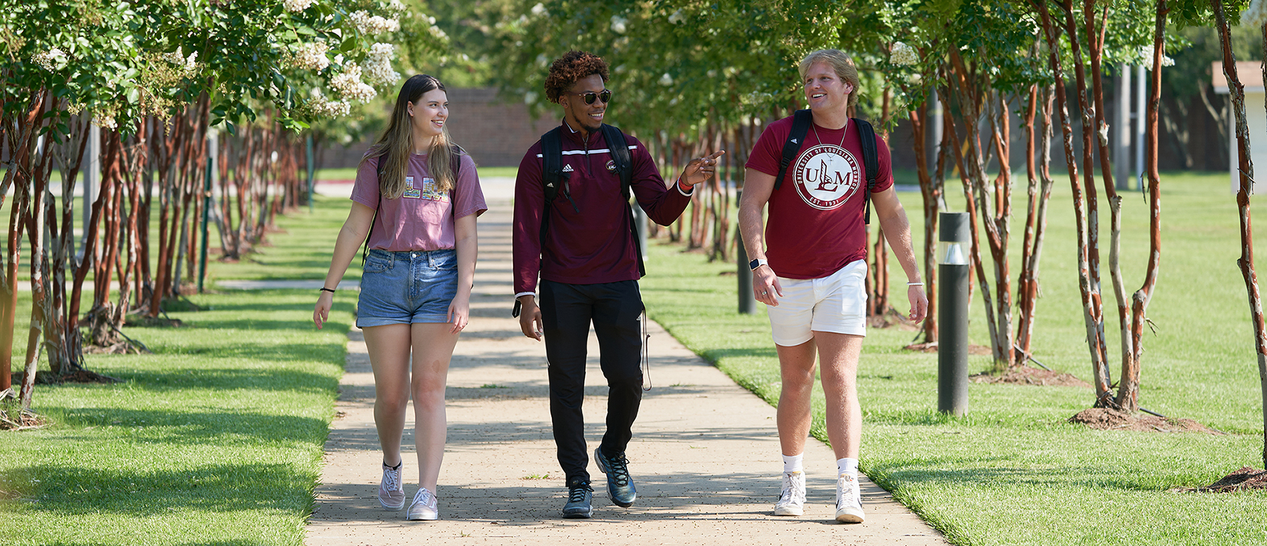 Three students walk along a sidewalk flanked by trees. The student in the middle points to his left. They are all smiling.