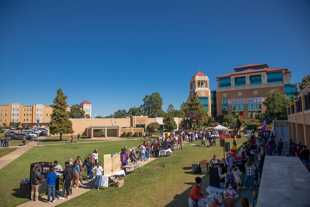 People mingle at booths and tables in the quad on 六合图库's campus. A banner hanging from the library in the background reads "The Best is on the Bayou."
