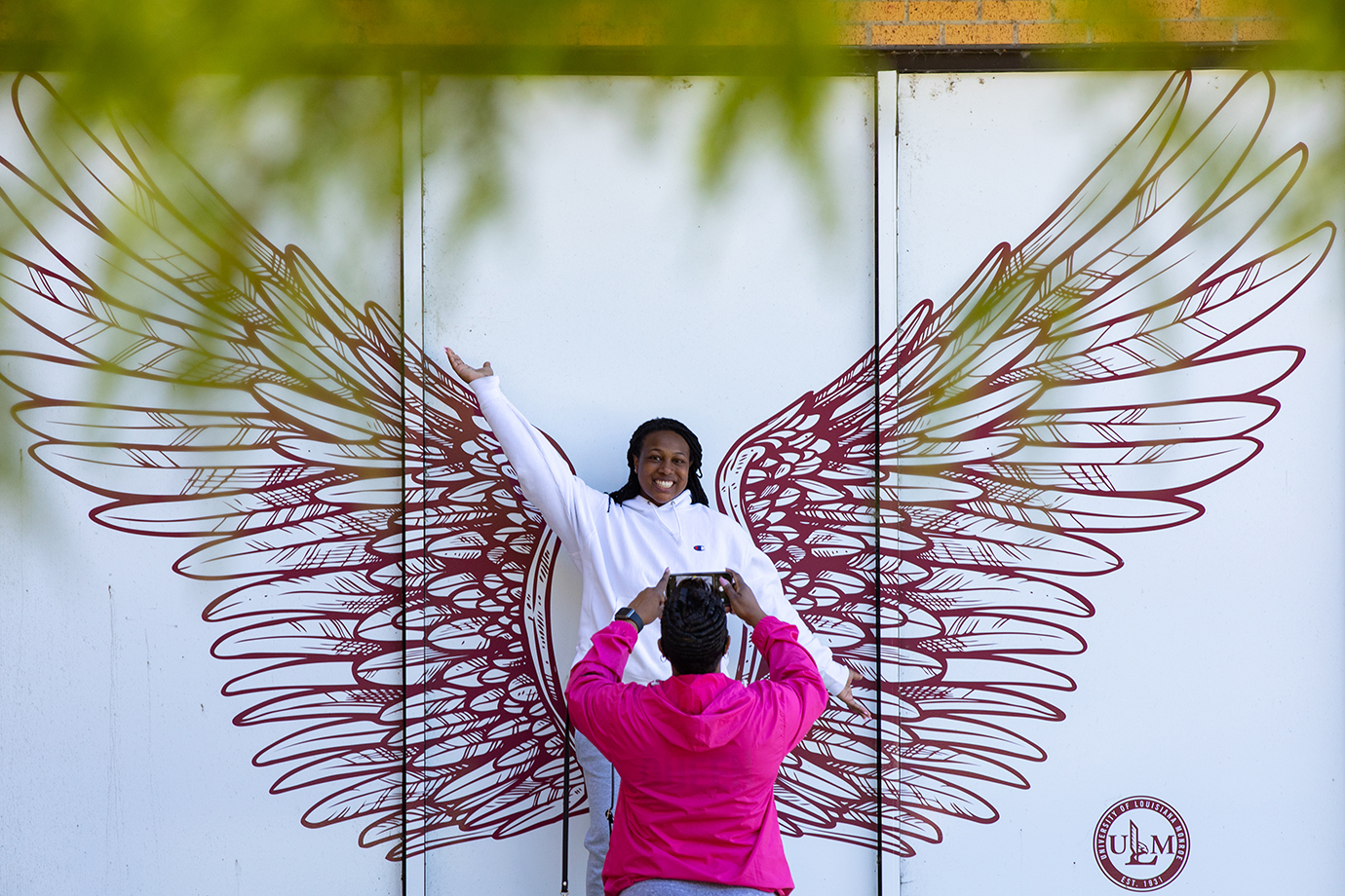 A student stands in front of a mural with wings. A woman takes her picture.